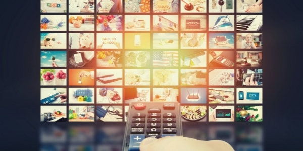 Evolucija TV oglaševanja - Roka z daljincem, usmerjena proti televizijskemu zaslonu z mnogimi vsebinskimi okni – simbol sodobnega TV okolja in evolucije TV oglaševanja, kjer ima gledalec več izbire in nadzora kot kdaj koli prej.