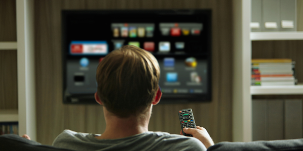 Oseba sedi na kavču in z daljincem upravlja pametni televizor, na katerem je prikazan uporabniški vmesnik z aplikacijami – simbolizira ciljano televizijsko oglaševanje (CTV).